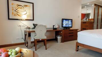 A hotel room with a bed, a desk with a computer, a chair, a TV, a trash bin, and a framed artwork on the wall.