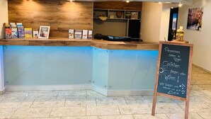 A reception desk with a wooden panel and a glass panel, a chalkboard with text, and a shelf with books and a basket. - Hotel Stadt Aurich (Aurich)