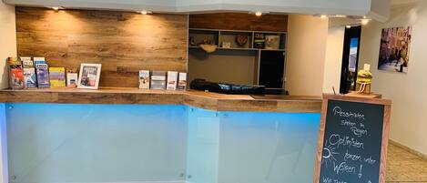 A reception desk with a wooden panel and a glass panel, a chalkboard with text, and a shelf with books and a basket.