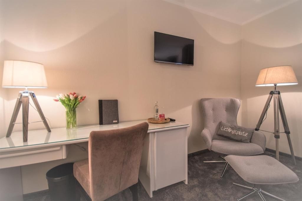 Una camera d'albergo con una scrivania, due poltrone, una televisione, una lampada e un tavolino con un vaso di fiori.