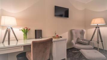 Una camera d'albergo con una scrivania, due poltrone, una televisione, una lampada e un tavolino con un vaso di fiori.