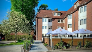 A brick building with a tiled roof, a paved walkway, and a tree with white flowers. - Hotel Stadt Aurich (Aurich)