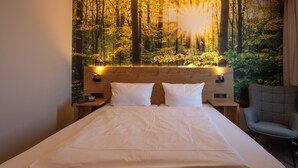 A hotel room with a bed, two pillows, a nightstand, a chair, and a wall mural of a forest. - Hotel Stadt Aurich (Aurich)