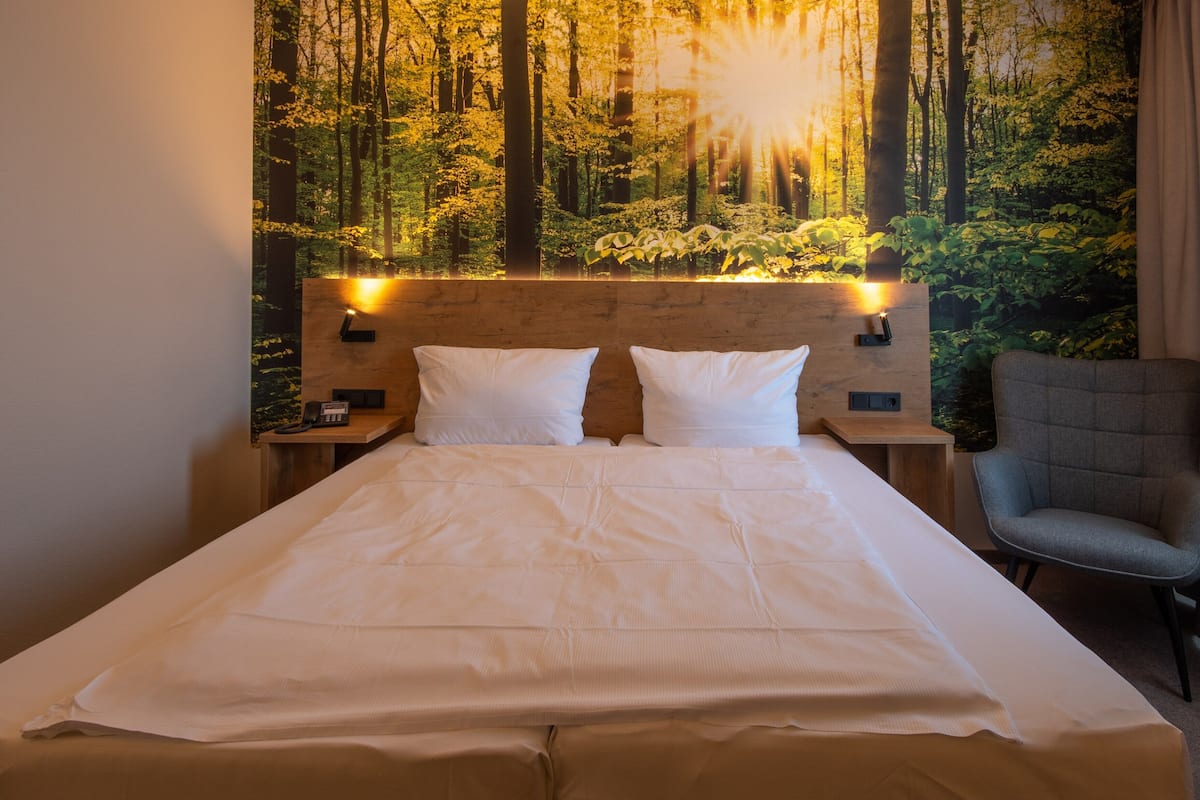 A hotel room with a bed, two pillows, a nightstand, a chair, and a wall mural of a forest.