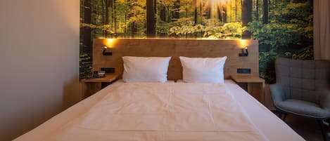 A hotel room with a bed, two pillows, a nightstand, a chair, and a wall mural of a forest.