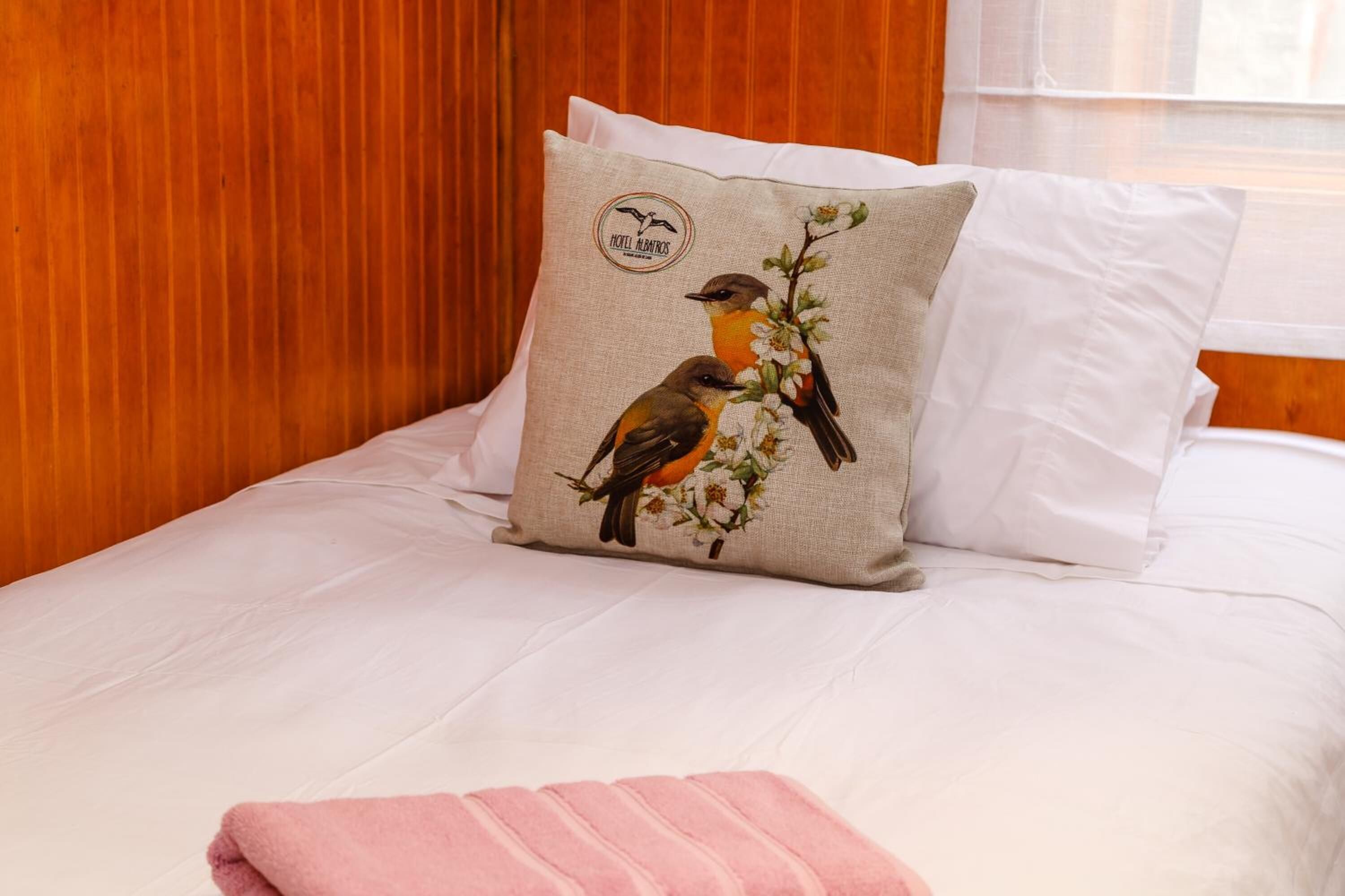 Una cama con almohada adornada con pájaros y flores, una colcha blanca y un cabecero de madera.
