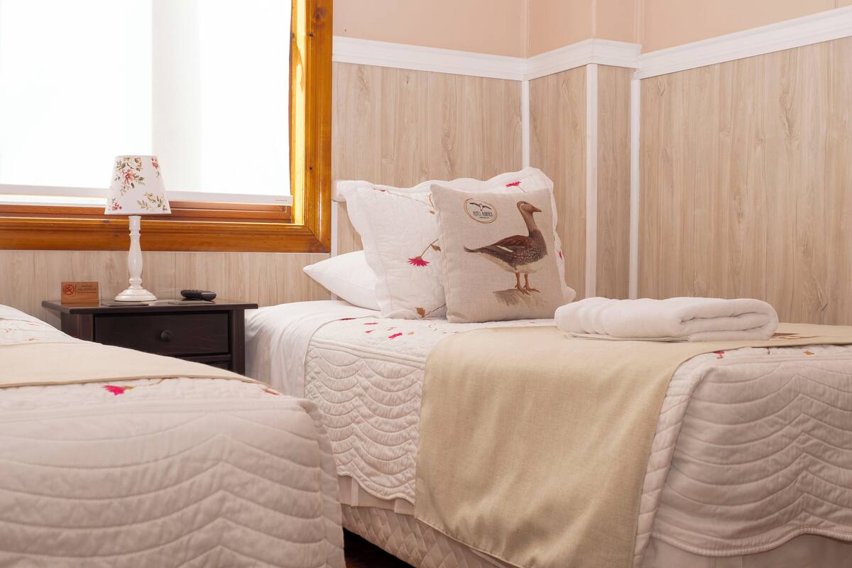 A hotel room with two beds, a nightstand, a lamp, and a window with a wooden frame.
