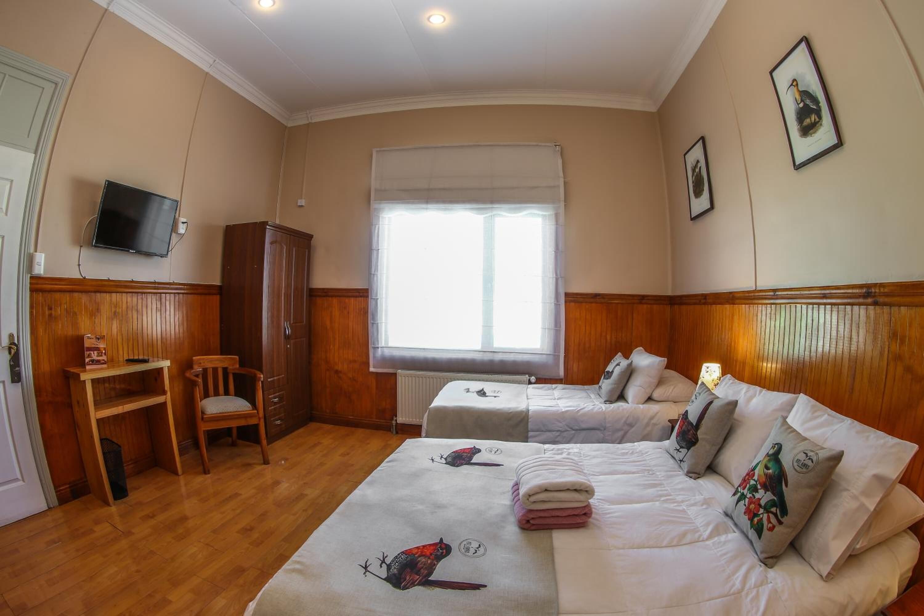 A hotel room with two beds, a television, a window with blinds, wooden paneling, and a small table with a chair.