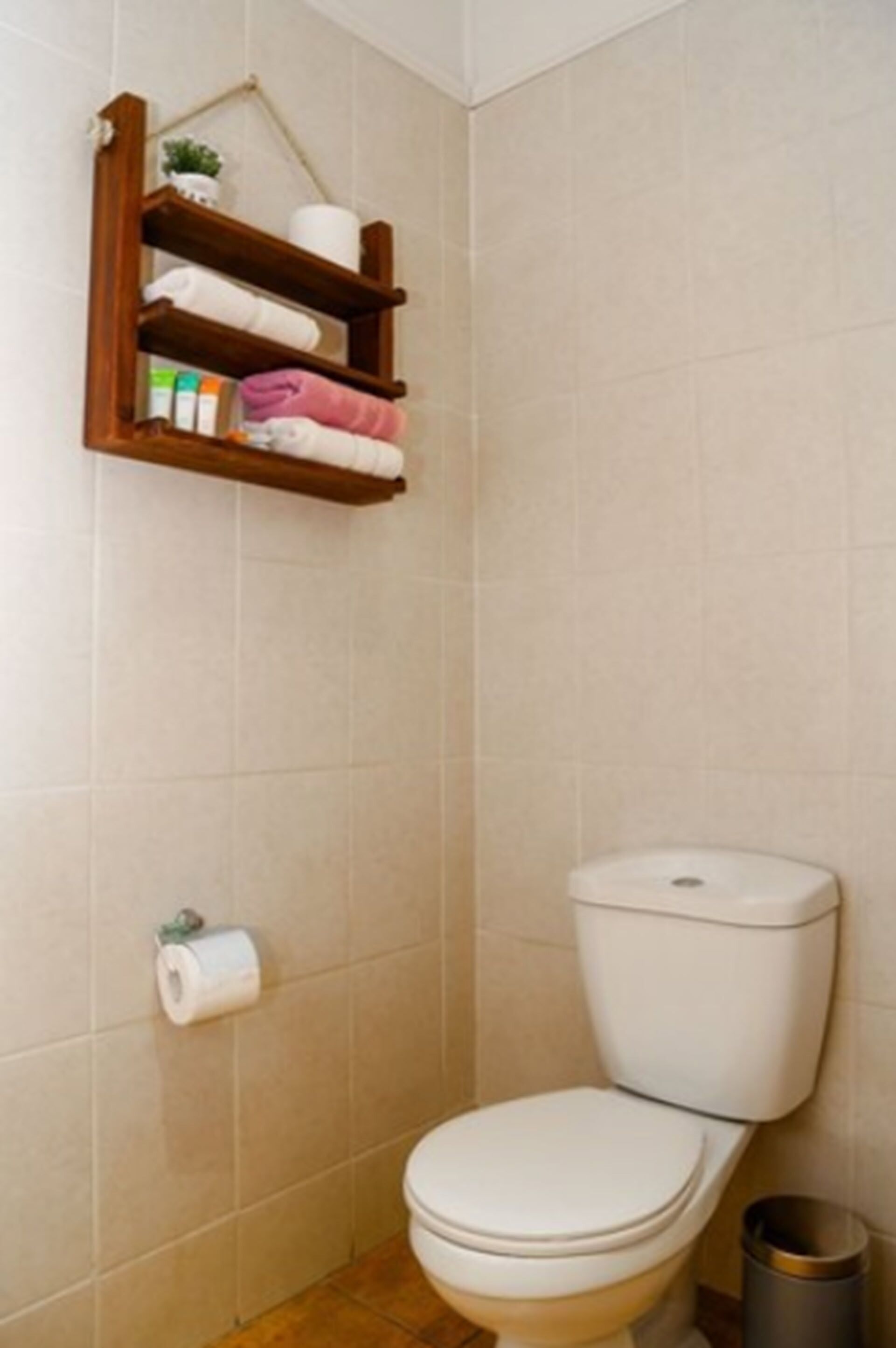 A small bathroom with a toilet, a shelf with toiletries, and a trash can.