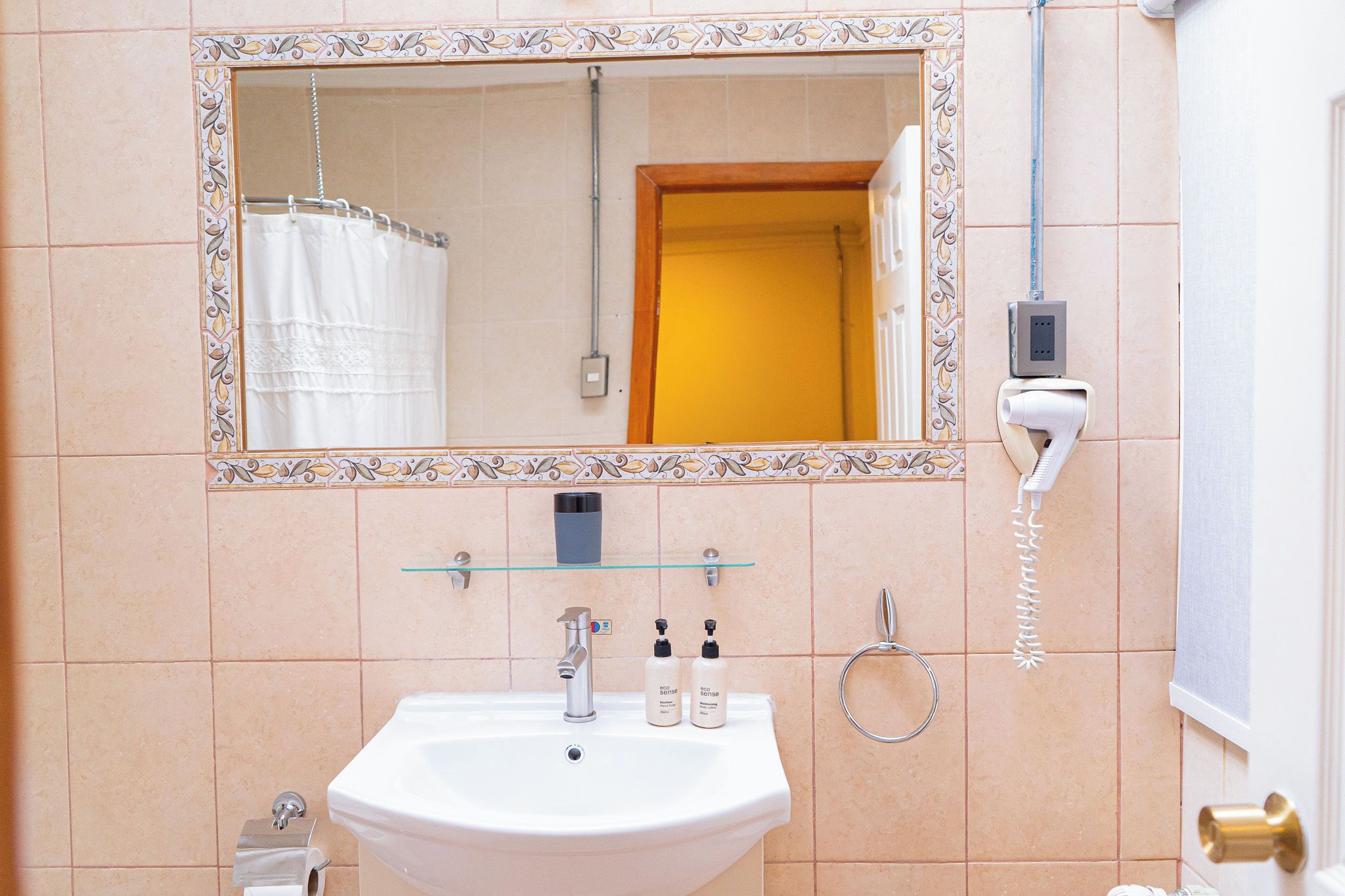 A bathroom with a white sink, a mirror with a decorative frame, a glass shelf, a hairdryer, and a towel rack.