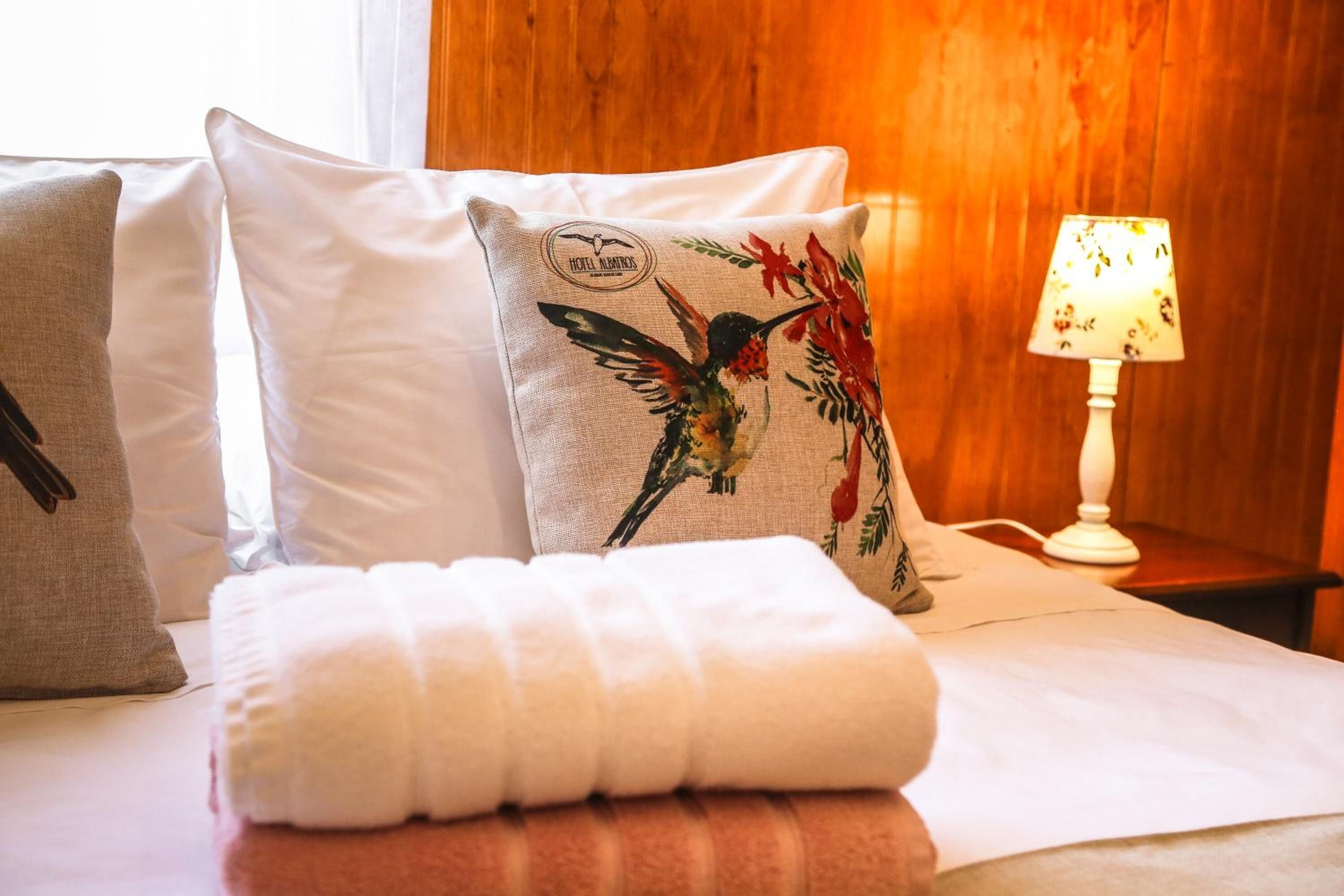 Una cama bien hecha con un almohadón blanco con diseño de pájaro, una toalla blanca y un cabecero de madera.