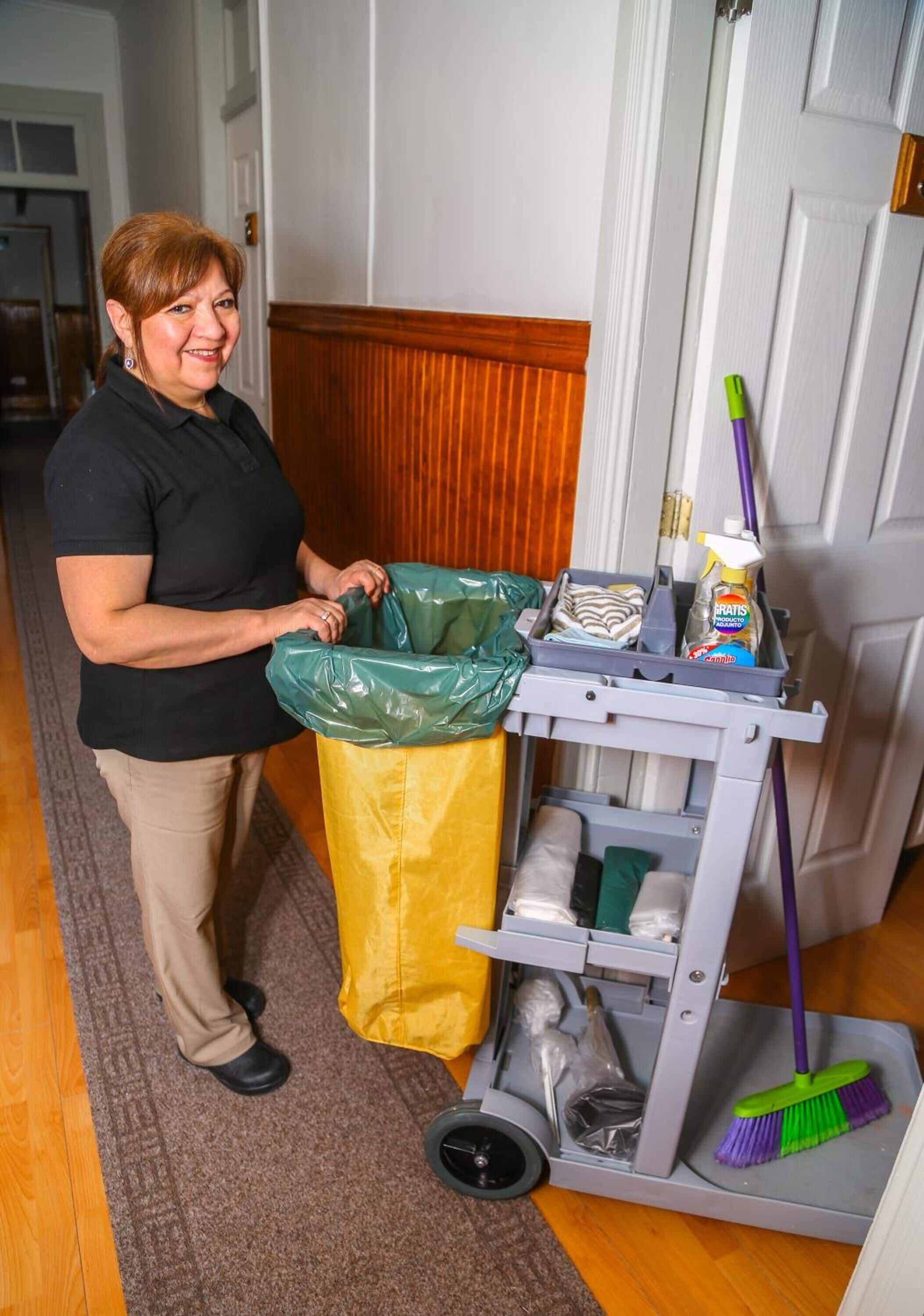 A person standing next to a cleaning cart with a trash bag, a broom, and cleaning supplies.