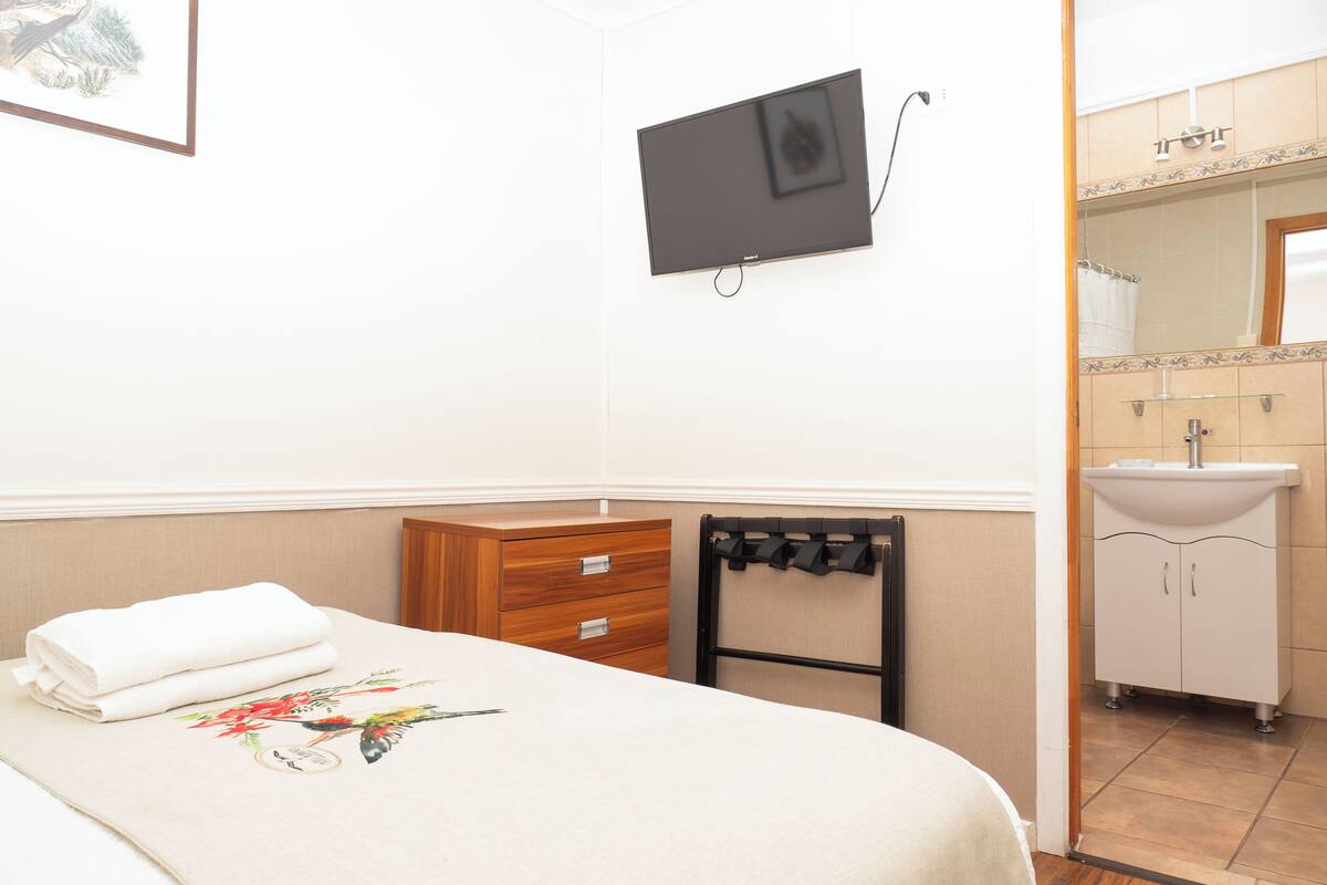 A hotel room with a bed, a TV mounted on the wall, a wooden dresser, and a bathroom with a sink and mirror.