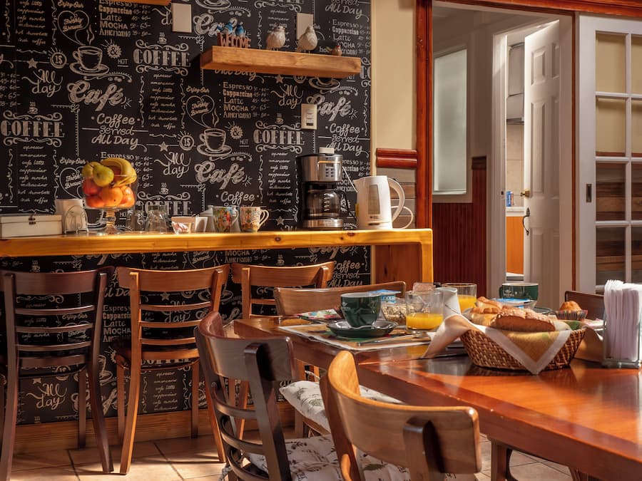Un café acogedor con una pared de pizarra que enumera los tipos de café y sus precios, muebles de madera y una mesa preparada para el desayuno.
