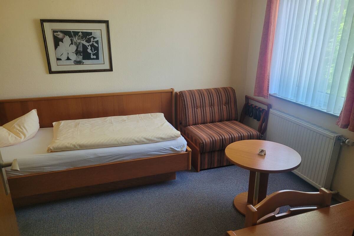 A compact hotel room with a single bed, a striped armchair, a small round table, and a radiator.
