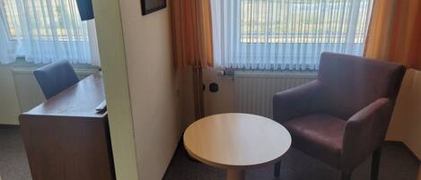 A hotel room with a desk, a round table, a chair, a lamp, and a window with curtains.