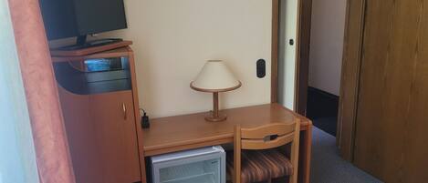 Habitación de hotel pequeña con un escritorio de madera, un refrigerador compacto, una lámpara, un televisor sobre un mueble y una puerta que da a otra habitación.