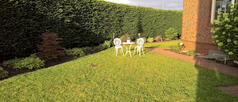 A well-maintained garden with a hedge, two white metal chairs, and a brick building.