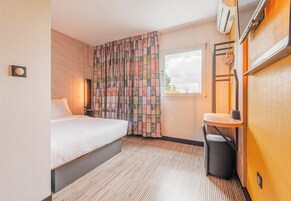 A hotel room with a bed, a small table, a chair, a window, and an air conditioning unit.