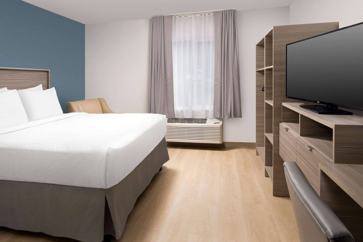 A modern hotel room with a large bed, a flat-screen TV, a built-in wardrobe, and a window with sheer curtains.