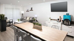 A modern hotel room with a wooden dining table, white chairs, a sofa, a TV, a coffee machine, and a potted plant. - WoodSpring Suites Seattle Redmond (Redmond)