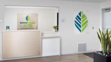 Uma área de recepção com balcão de madeira, sinalização da WoodSpring Suites, um vaso de planta e uma obra de arte fixada na parede.