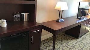 A hotel room with a wooden desk, a microwave, a lamp, a water bottle, a coffee maker, and a TV.