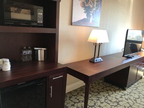 A hotel room with a wooden desk, a microwave, a lamp, a water bottle, a coffee maker, and a TV.