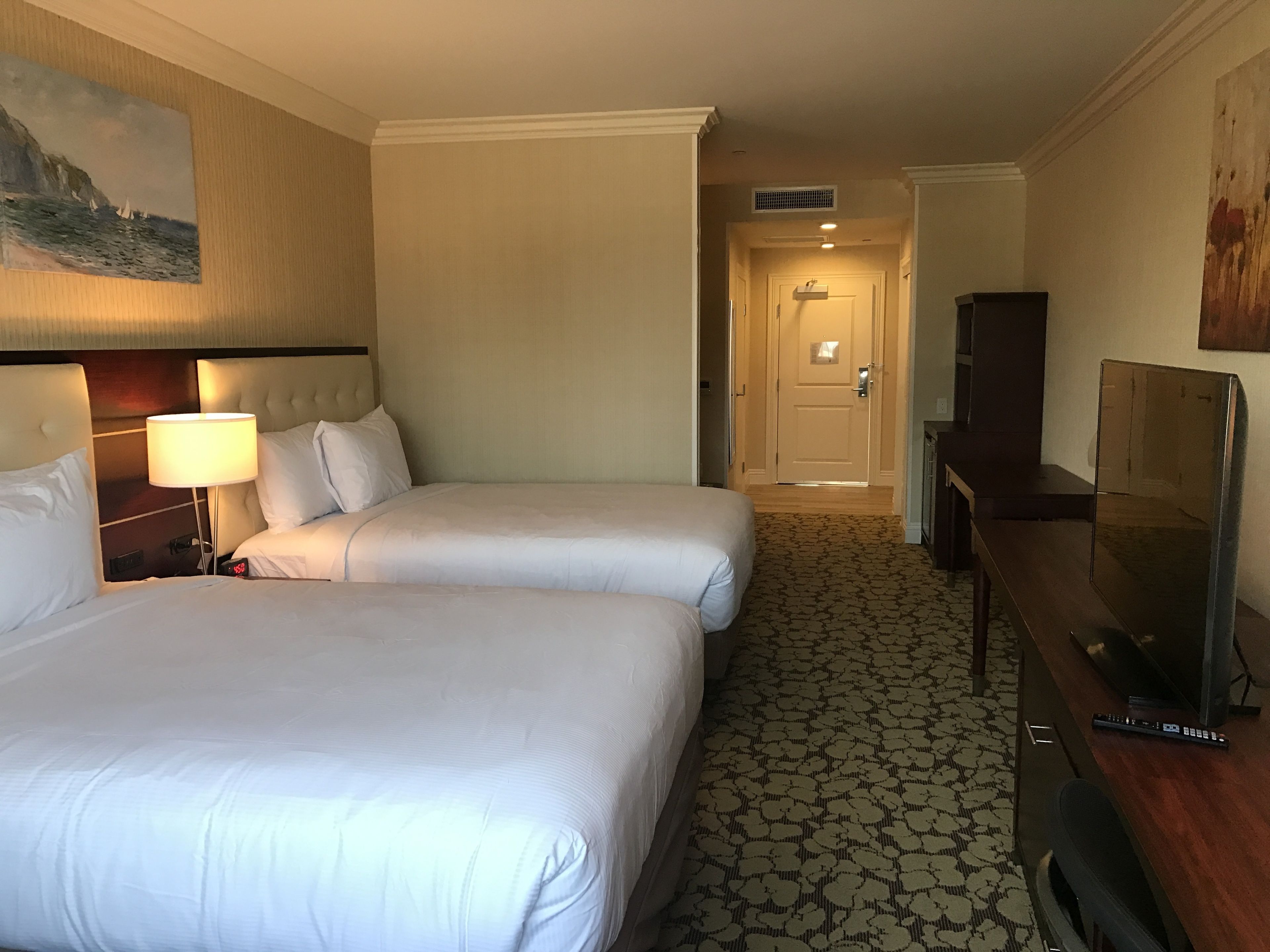 A hotel room with two beds, a desk with a television, and a painting on the wall.