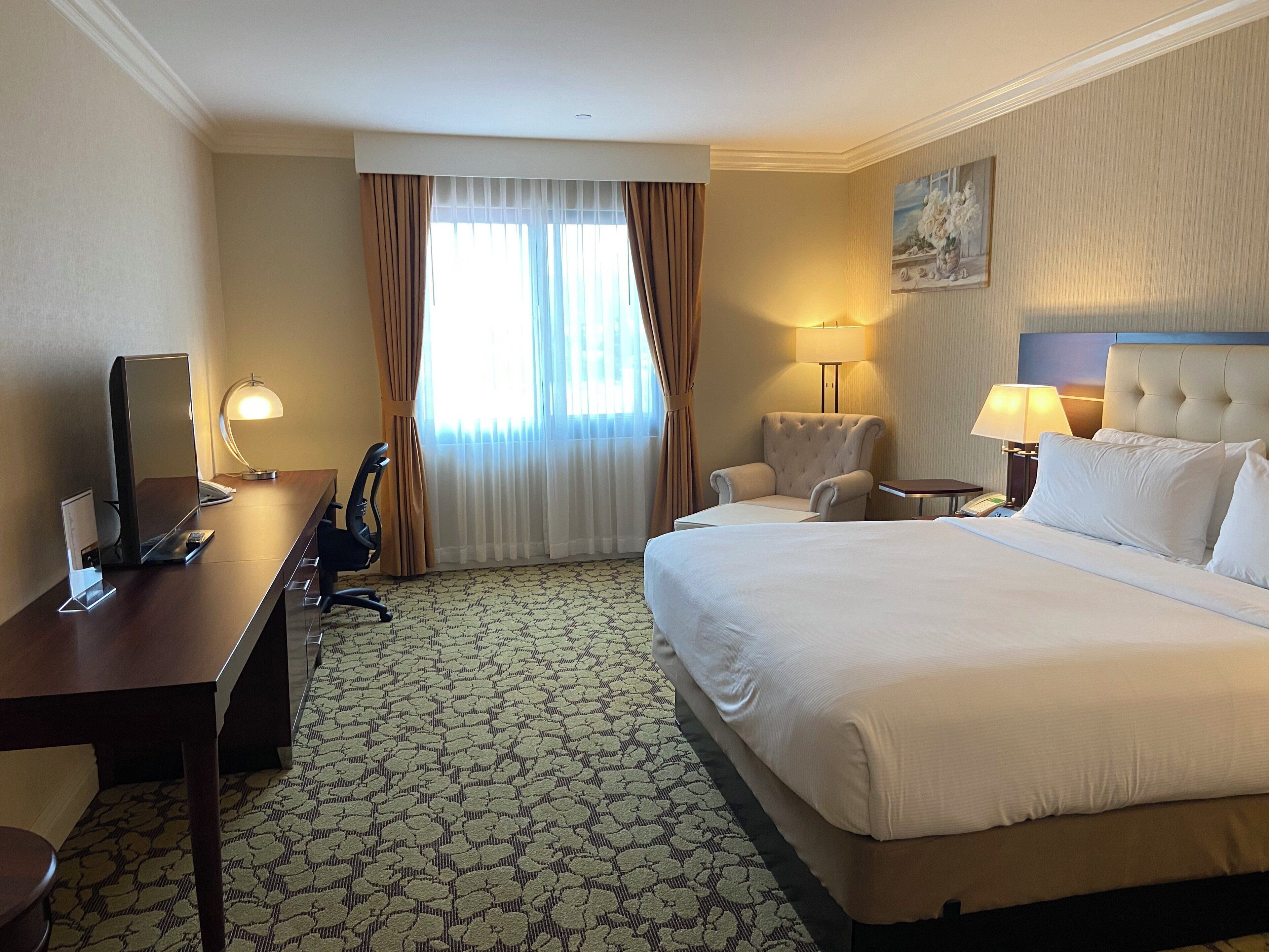 Una habitación de hotel con una cama, un escritorio con una computadora, una silla, una lámpara y un cuadro en la pared.
