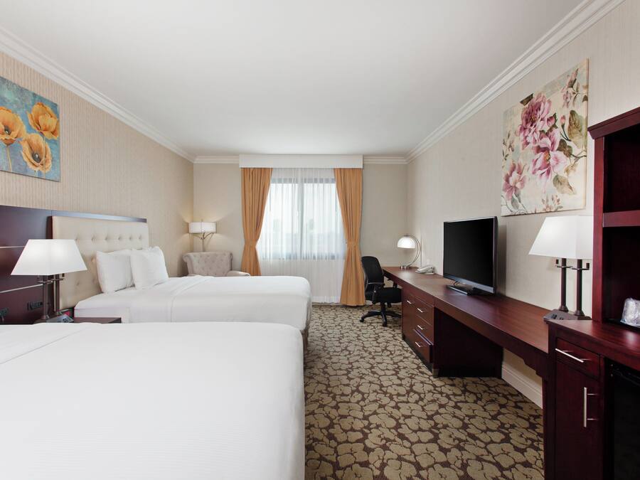 Habitación de hotel con dos camas, un escritorio con televisor y un ventanal grande con cortinas.