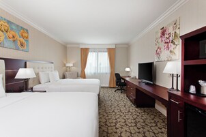 Quarto de hotel com duas camas, uma mesa de trabalho com televisão e uma janela grande com cortinas.