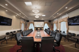 Un salón de conferencias con una larga mesa, sillas de cuero negro, una pantalla de proyector y dos televisores en las paredes.