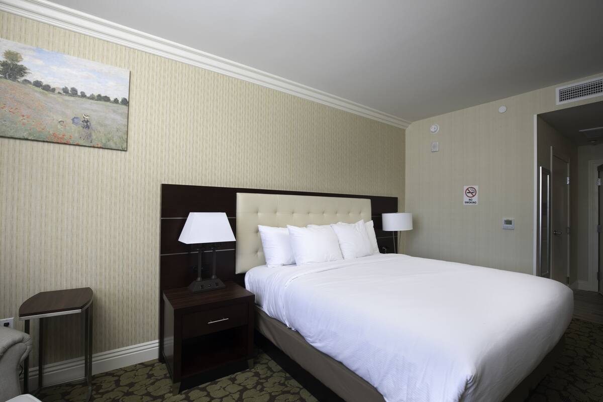 Une chambre d’hôtel avec un grand lit, deux tables de chevet avec des lampes, un tableau au mur et une porte sur la droite.