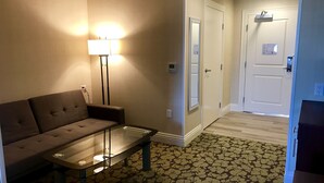 A hotel room with a sofa, a glass-top table, a patterned carpet, a hallway with a door, and a wall-mounted light fixture.