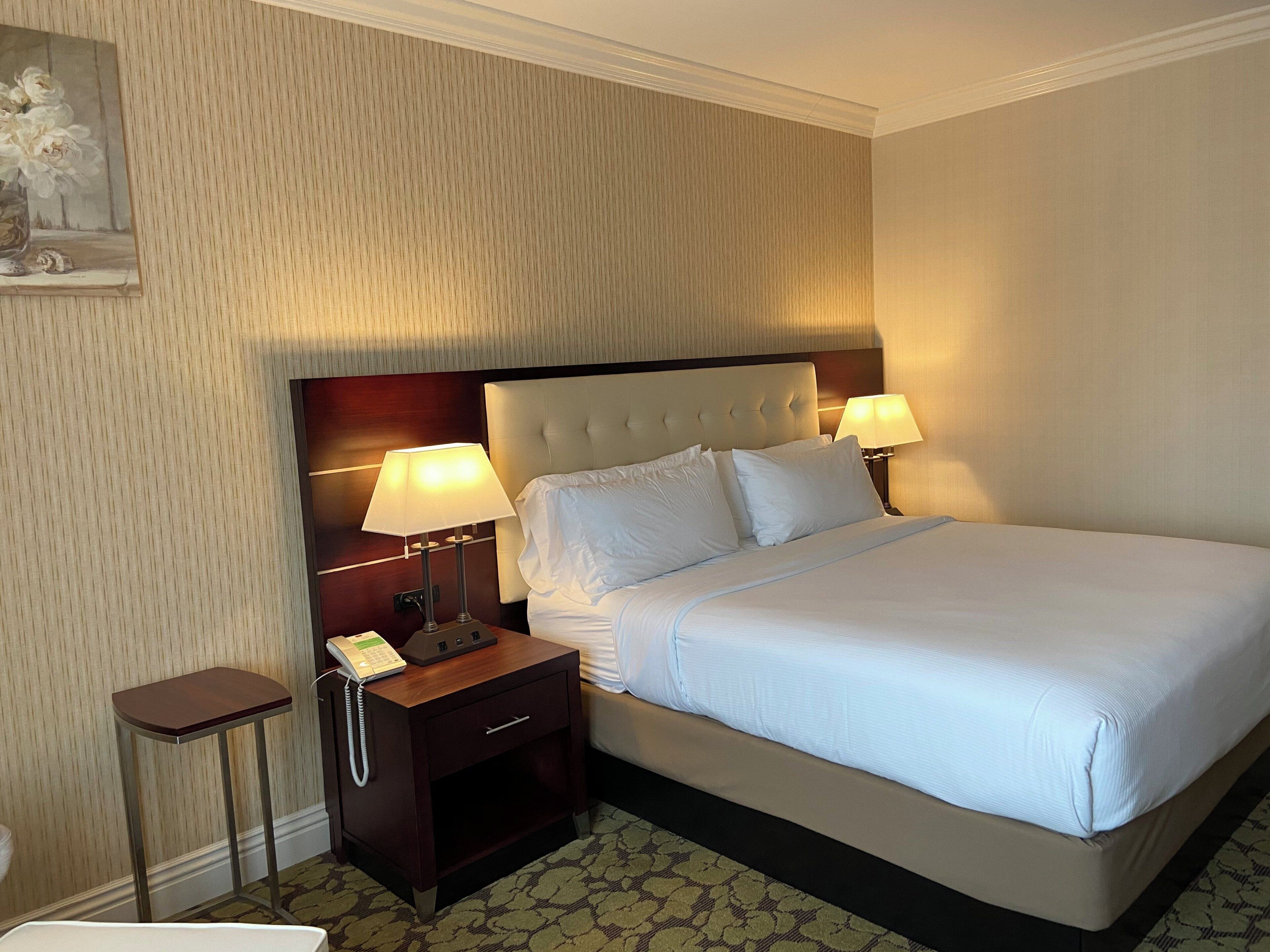Una habitación de hotel con una cama grande, dos lámparas de noche, una mesita de noche con teléfono y una alfombra estampada.