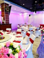 A banquet hall with round tables, white chairs with red sashes, and floral centerpieces.