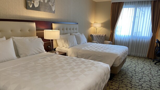 A hotel room with two beds, a headboard, a nightstand, a lamp, a chair, and a window with curtains.