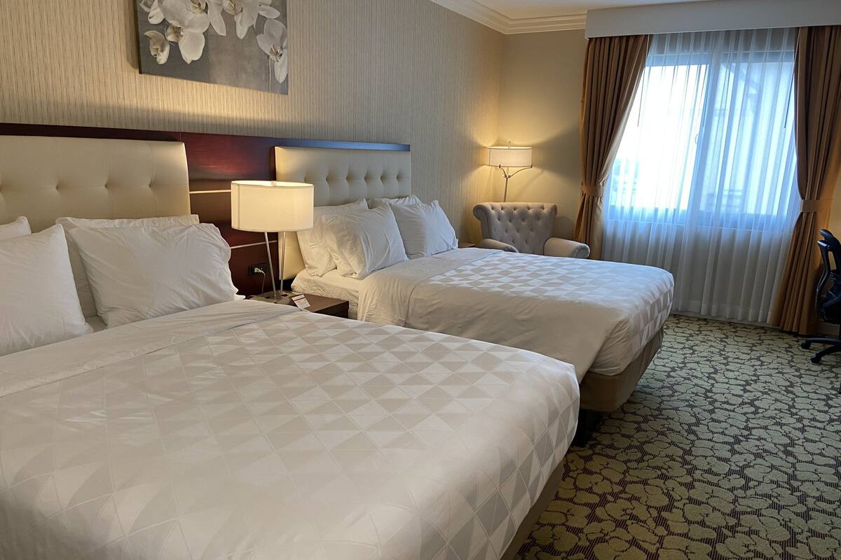 Une chambre d’hôtel avec deux lits, une tête de lit, une table de chevet, une lampe, une chaise et une fenêtre avec des rideaux.