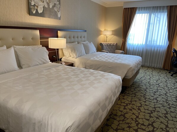 A hotel room with two beds, a headboard, a nightstand, a lamp, a chair, and a window with curtains.