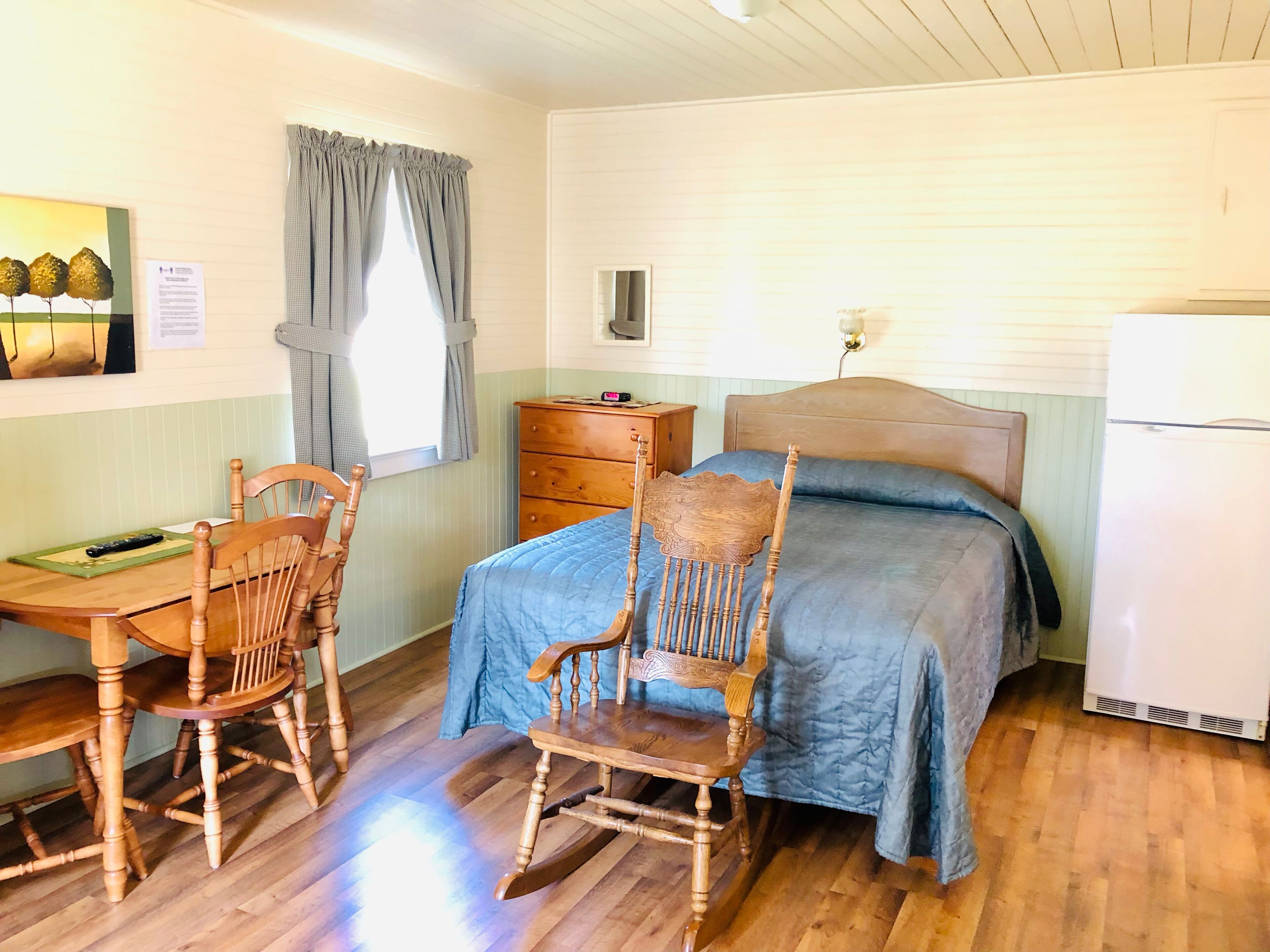 A cozy room with a bed, a wooden desk with a chair, a dresser, and a window with curtains.