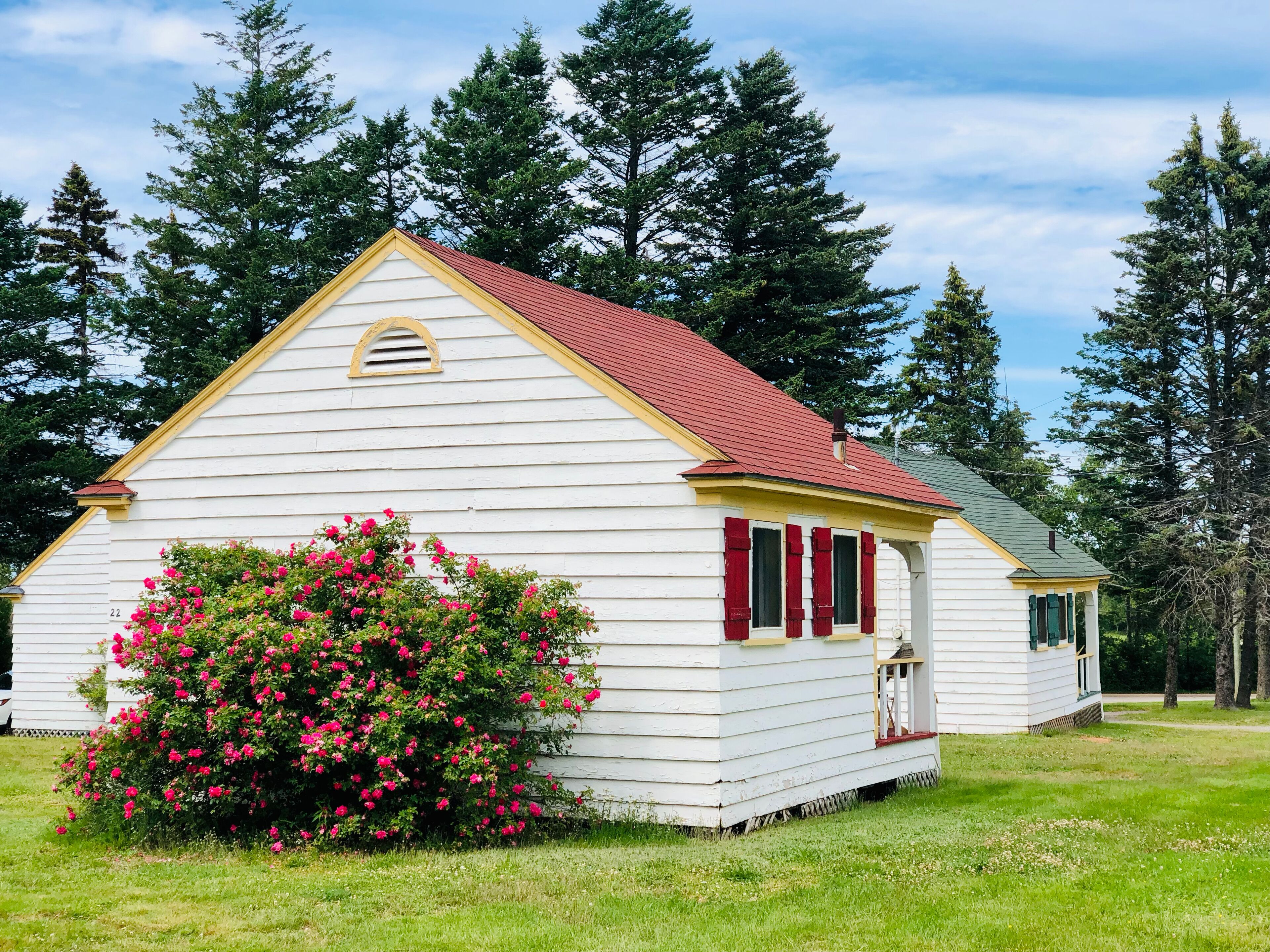 A white building with a red roof, surrounded by green trees and a bush with pink flowers.