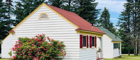 A white building with a red roof, surrounded by green trees and a bush with pink flowers.