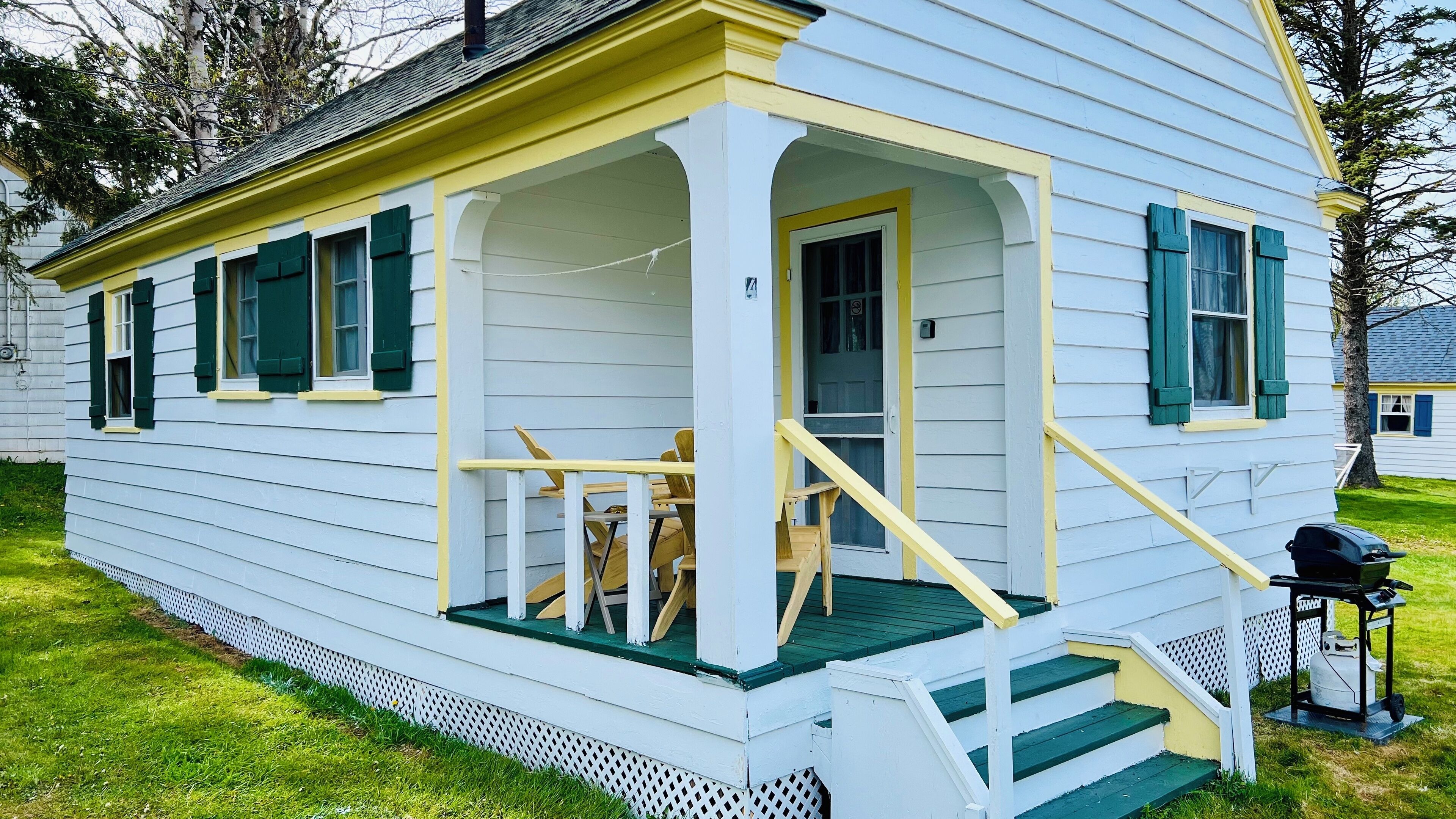 A white house with green shutters, a porch with wooden chairs, and a small staircase leading to the entrance.