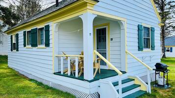 A white house with green shutters, a porch with wooden chairs, and a small staircase leading to the entrance.