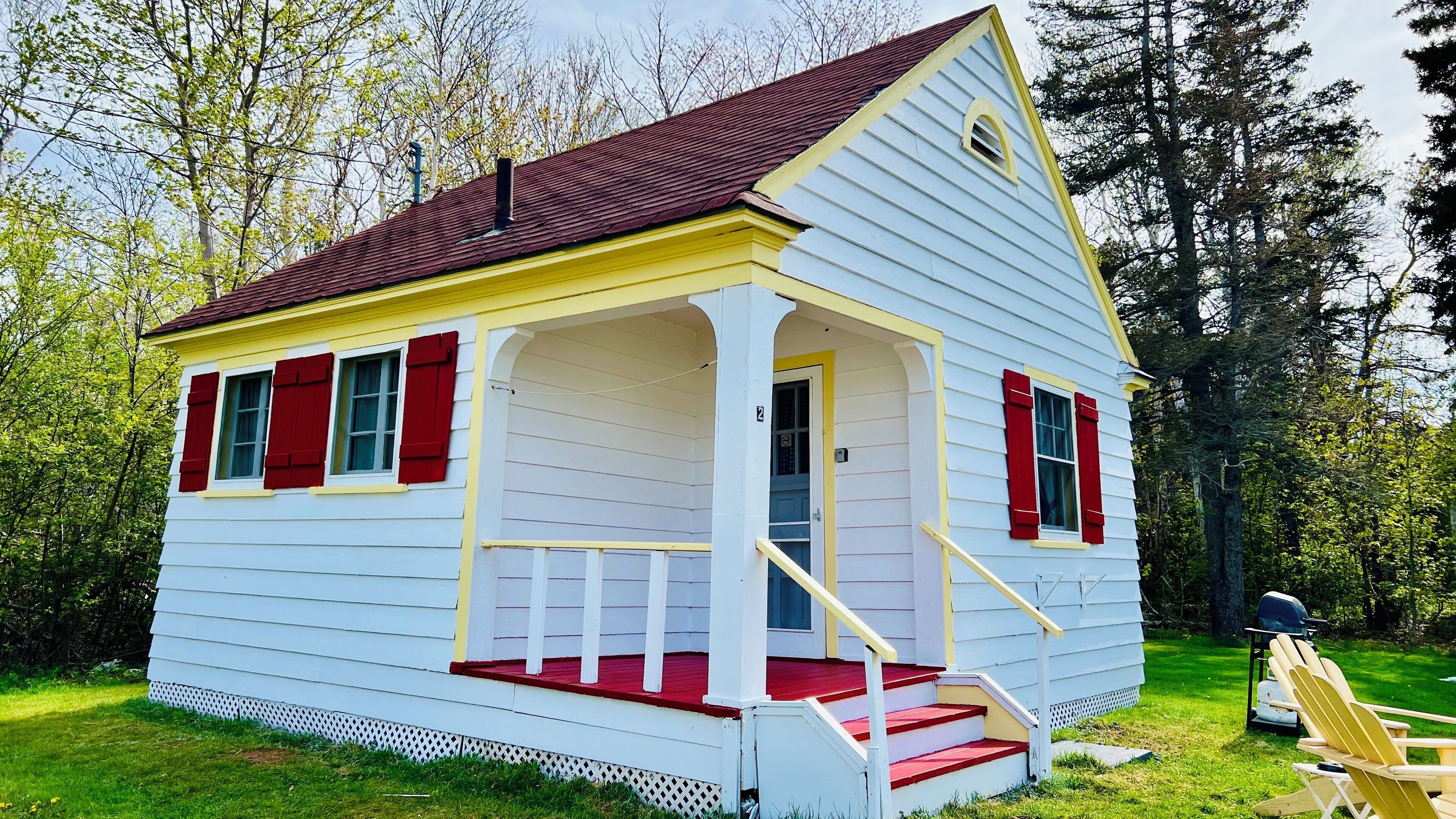 A small, single-story house with a porch and red shutters, surrounded by trees.