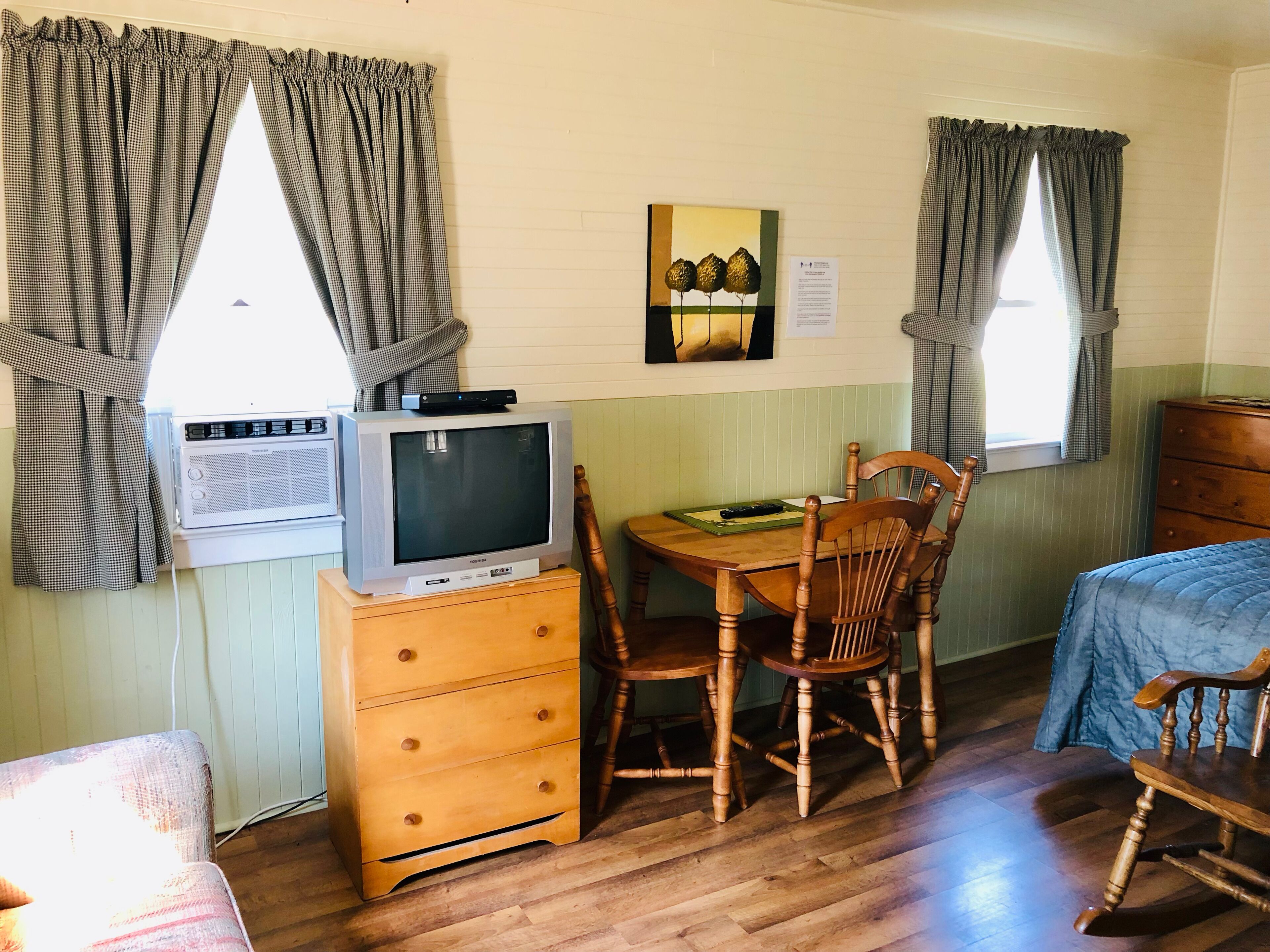 A room with a wooden table, two wooden chairs, a television on a dresser, and a bed with a blue blanket.