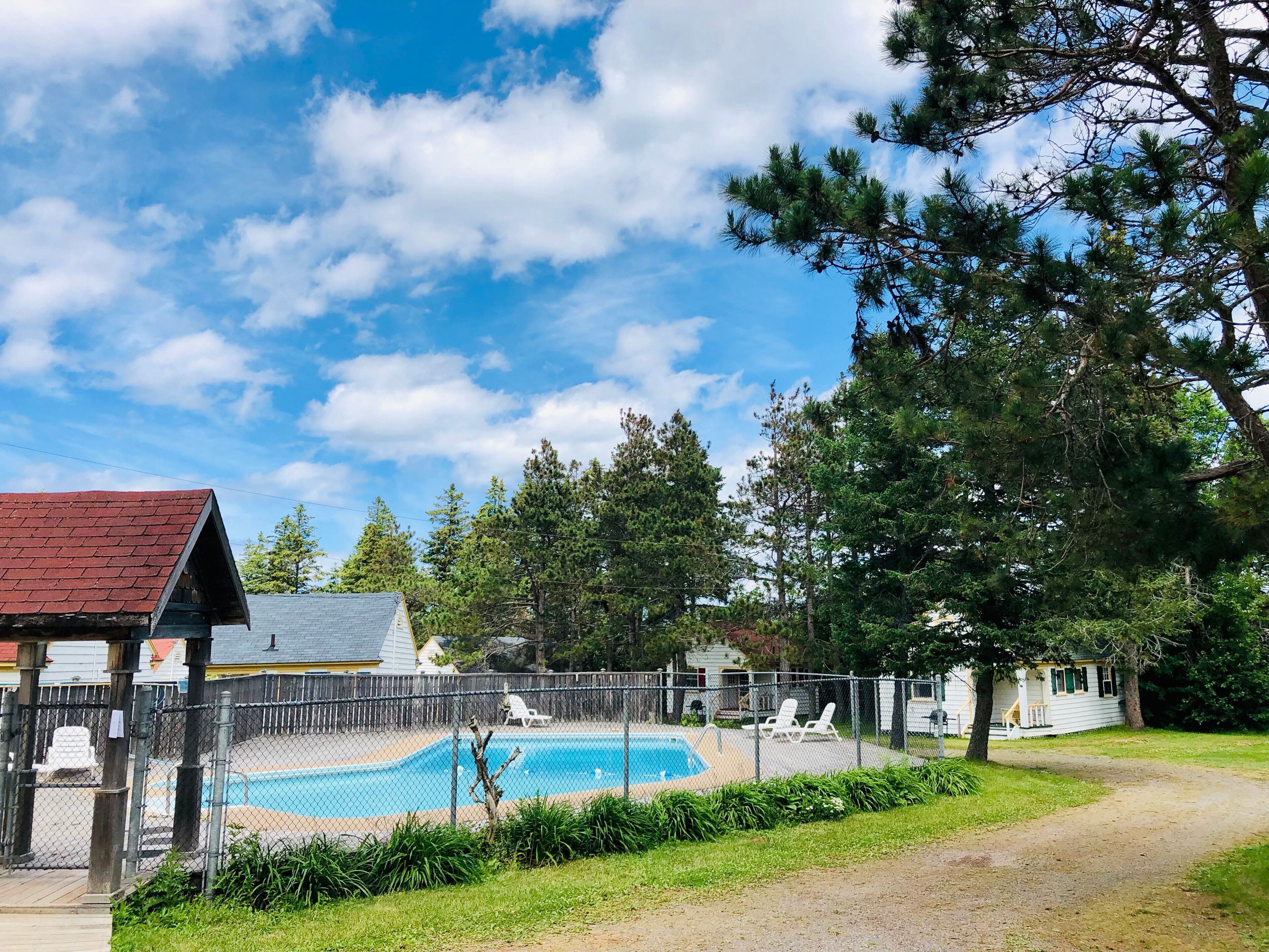 Une piscine entourée d’une clôture, avec une terrasse en bois, le tout bordé de verdure et surplombé par un ciel dégagé.