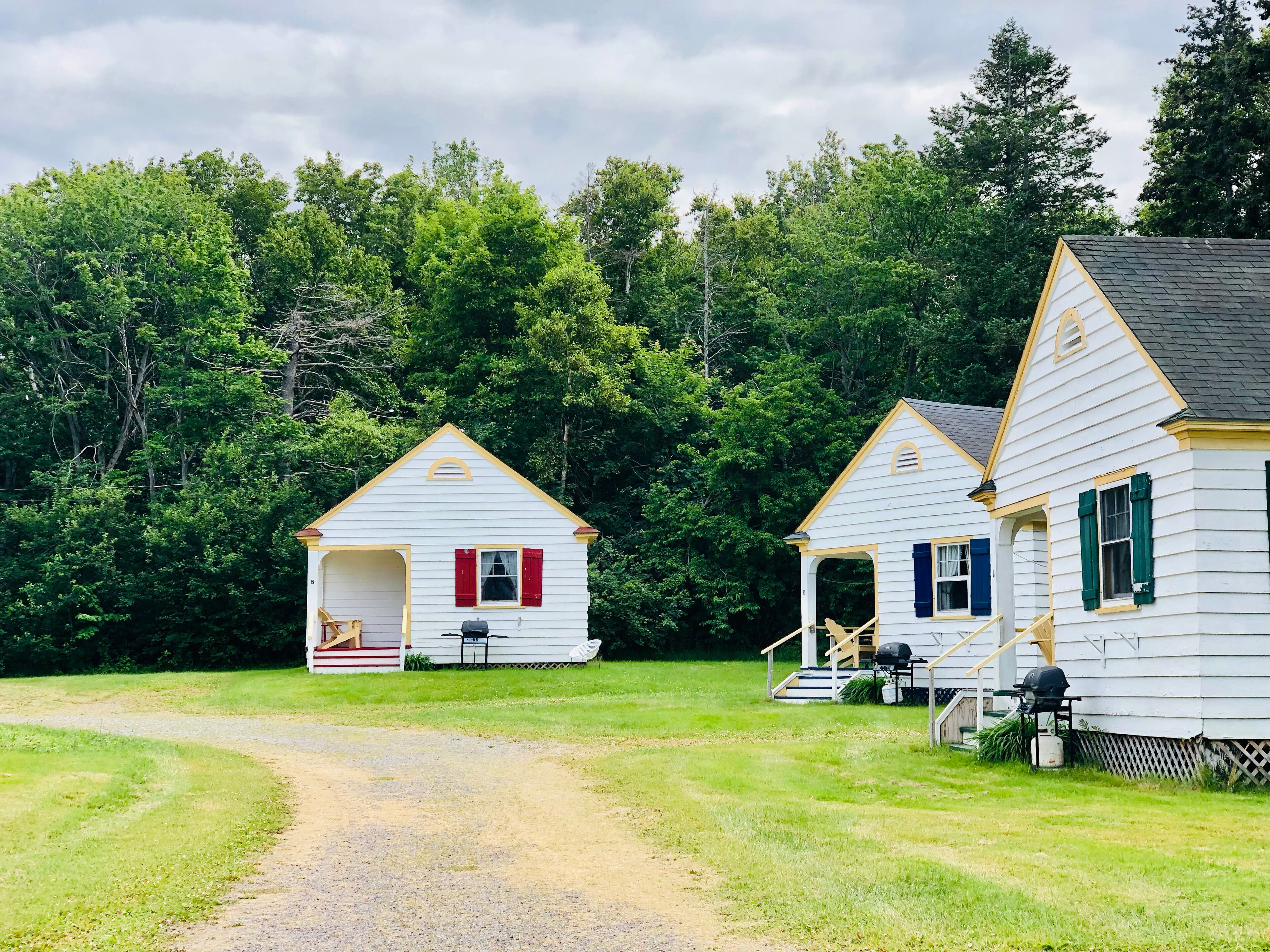 Two white cabins with red shutters, a gravel path leading to them, and a forested backdrop.