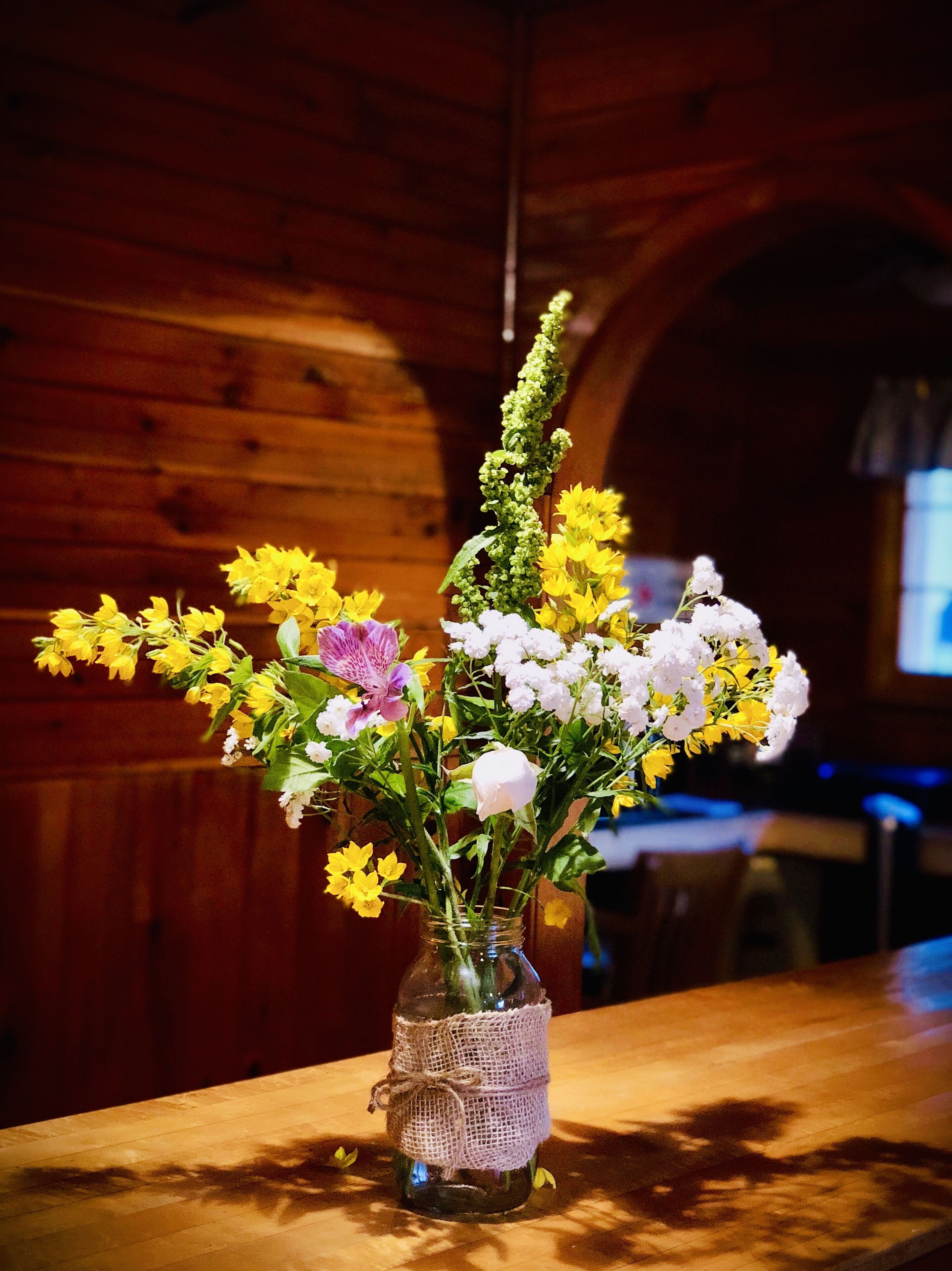 Un florero de vidrio con envoltorio de arpillera, lleno de flores amarillas y blancas, sobre una mesa de madera en una habitación con paredes de madera y un arco visible.